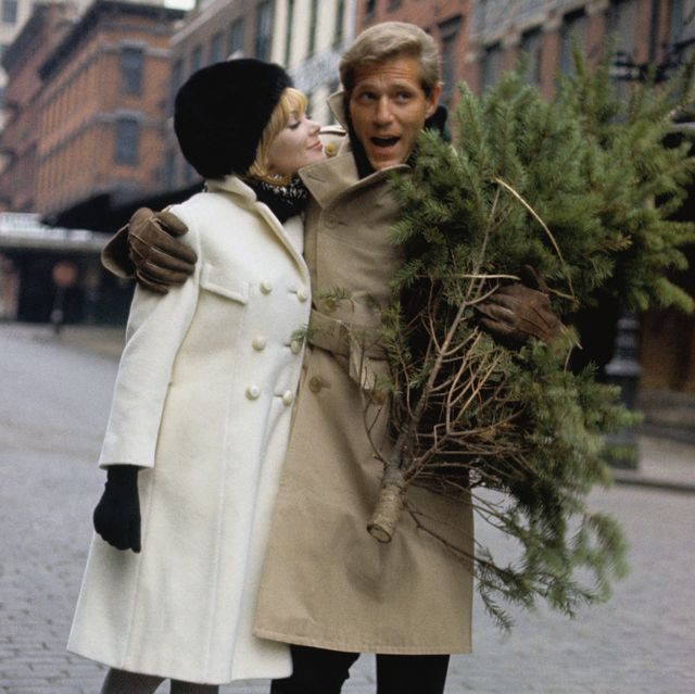 george segal and maggie london walk down a city street, their arms wrapped around each other, he carries a small christmas tree she wears a white wool coat by dani jrs, black and white muffler by einier accessories, black bubble hat by veaumont, black gloves by hansen, and grey stockings by berkshire he wears a khaki trench coat with slacks photo by louis faurercondé nast via getty images