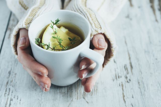 human hands holding cup of tea with thyme herb and lemon slices on a wooden table,