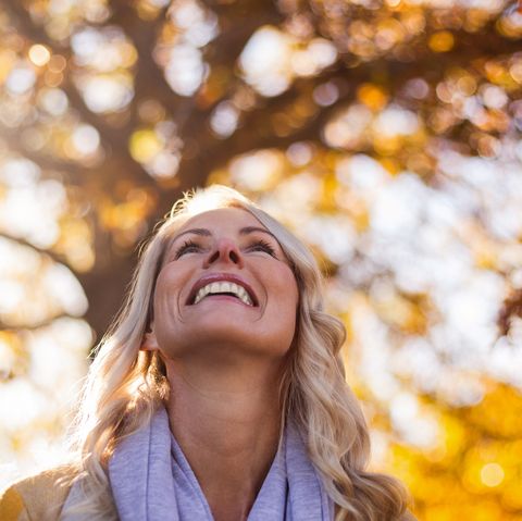 smiling woman looking up against trees at park during autumn
