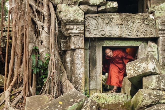 cambodian monk ducks under a short doorway to explore an ancient temple with fallen stones and tangled roots encroaching the structure, ta prohm, angkor wat, siem reap, cambodia, asia