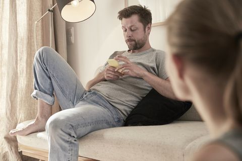 Man at home sitting on couch looking at cell phone with women in foreground