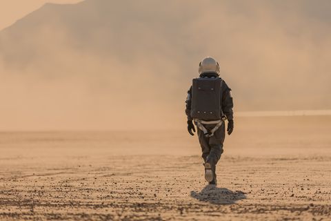 full length photo of astronaut in space suit and helmet on mars walking away from the camera towards a dust storm in the distance, red orange all around