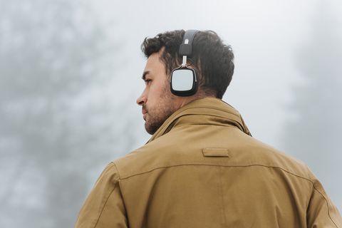 Horizontal portrait of young Caucasian man walking in foggy mountain, listening to music outside. Rear view of Caucasian man wearing trendy coat and bluetooth headphones. People, technology concept.