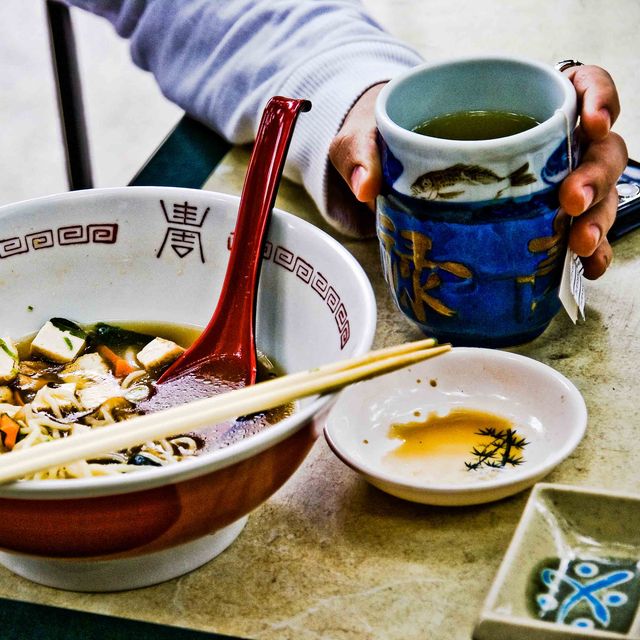young man at outdoor restaurant table eating large bowl of japanese noodles with tofu and vegetables cup of green tea, chopsticks and condiment dishes on table