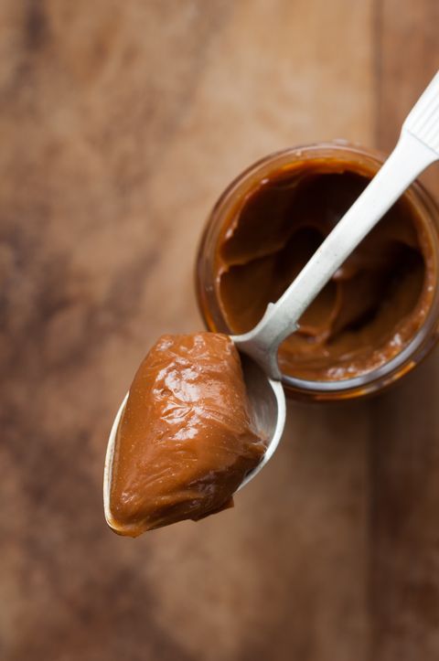 close up of a spoon with dulce de leche on top of a glass jar with wooden background