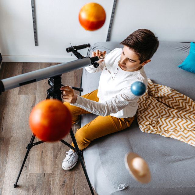 boy sitting on bed looking into telescope with planet mobile above his head