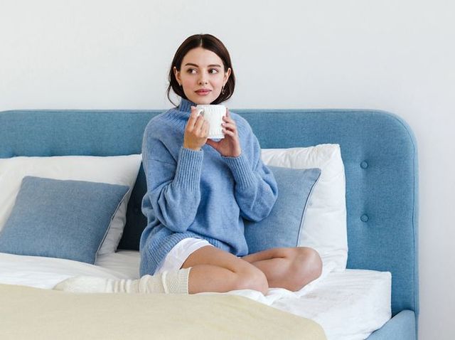 Girl in a blue sweater in interior Hygge style with a cup of hot tea in her hands sits on the bed