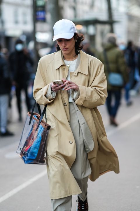 a woman wearing a trench coat, white tshirt, khaki pants, white baseball cap, tote bag, and sneakers on the street