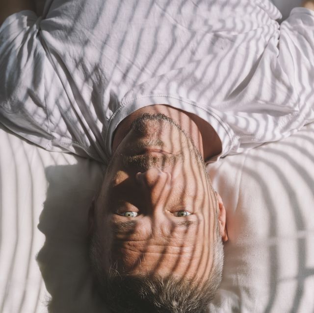 handsome adult man wakes up in the morning, lying on back, with sunlight illuminated him through window with blinds
