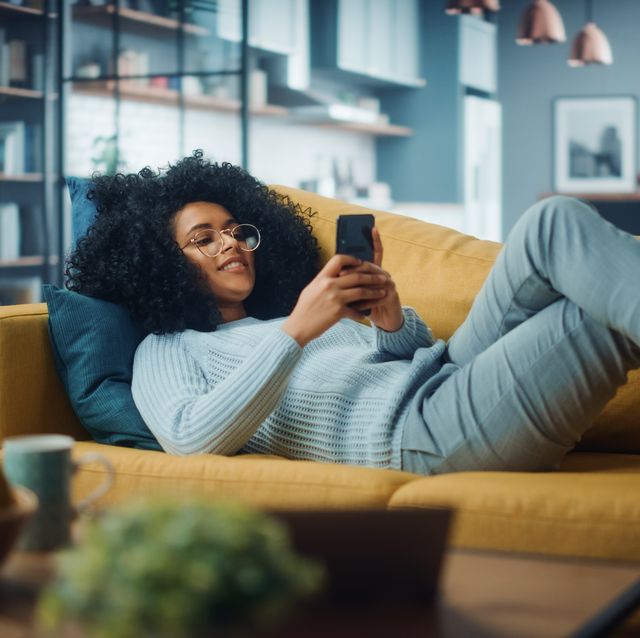 happy beautiful latina female using smartphone in cozy living room at home female resting on comfortable sofa she's browsing the internet and checking videos on social networks and having fun