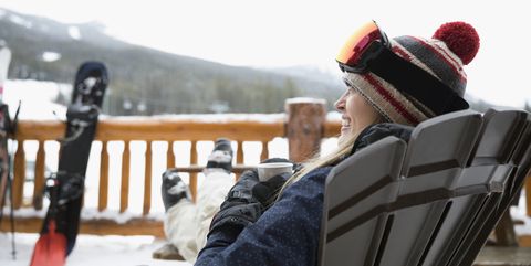 Happy female snowboarder relaxing apres-ski with coffee on snowy ski resort balcony