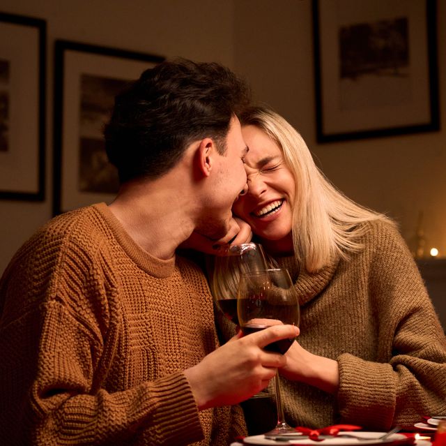happy young couple in love hugging, laughing, drinking wine, enjoying talking, having fun together celebrating valentines day dining at home, having romantic dinner date with candles sitting at table