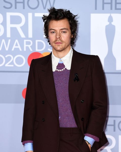 harry styles poses on the red carpet, wearing a black suit over a purple sweater, a blue button up with a white lace collar, and a pearl necklace