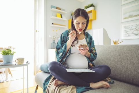 una chica embarazada toma un desayuno compuesto por cereales y leche en el sofá de su salón