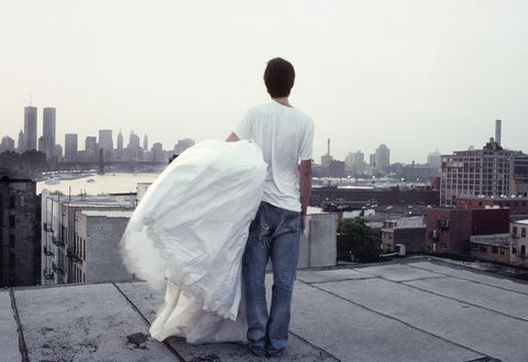person wearing white t shirt and jeans and carrying a poofy white dress stands on a rooftop looking at the manhattan skyline, including the twin towers