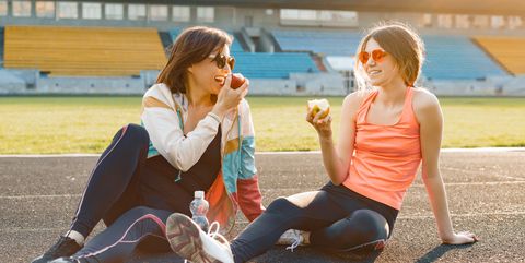 smiling fitness mother and teen daughter together eating apple sitting on stadium after training on sunny spring day