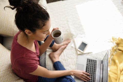 Hispanic woman sitting on bed drinking tea and using laptop