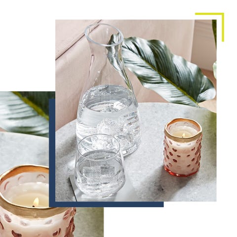 a burning candle sitting on a marble side table alongside a water jug with glasses