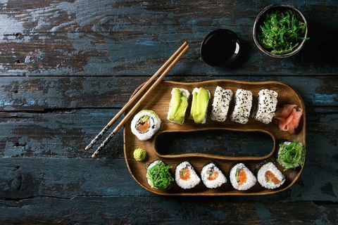 Homemade sushi rolls set with salmon, sesame seeds and avocado serving in wood plate with pink pickled ginger, soy sauce, wasabi, seaweed salad, chopsticks over dark wooden background. Top view, space