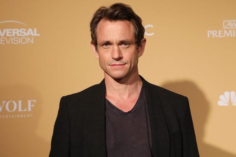 Hugh Dancy hugh dancy, a man stands looking at the camera, brown hair in loose waves around head, black tshirt and jacket