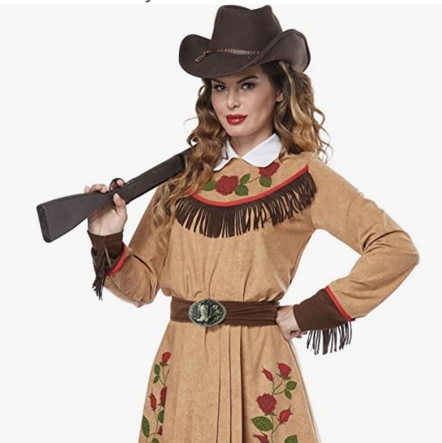 woman dressed as annie oakley in tan dress with roses on chest and sides of skirt and fringe on cuffs and across chest she is wearing a brown cowboy hat and holding a faux rifle on her shoulder