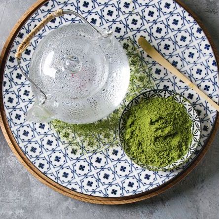 ingredients for making matcha ice drink green tea matcha powder in ceramic bowl, traditional bamboo spoon, whisk on plate, glass teapot, ice cubes over grey texture background flat lay, space