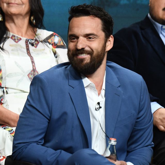 jake johnson in a blue suit and white shirt smiling while sitting down on stage on a press tour