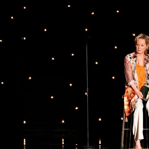 jean smart wearing an orange shawl and white pants sitting on a stage in her new show hacks