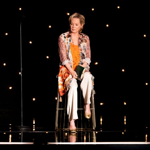 jean smart wearing an orange shawl and white pants sitting on a stage in her new show hacks