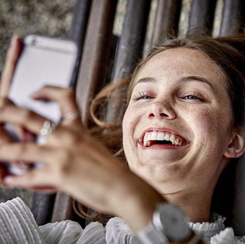 Laughing young woman lying on a bench using cell phone