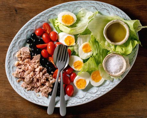 layered french nicoise salad with canned tuna fish, olives and eggs, served with olive oil dressing and sea salt on ceramic plate over wooden background, flat lay, copy space