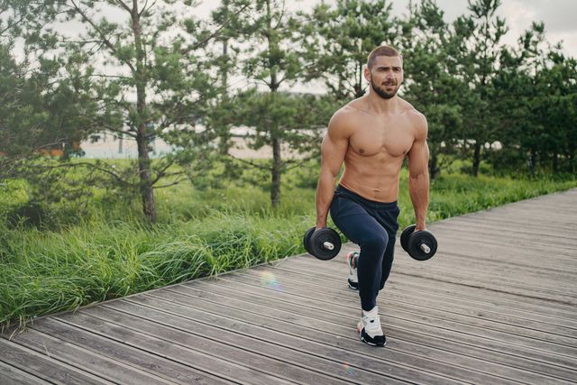 motivated bearded man with naked torso makes squat with heavy barbells, does physical exercises outdoor, enjoys workout, poses near green trees, doing weightlifting healthy lifestyle concept