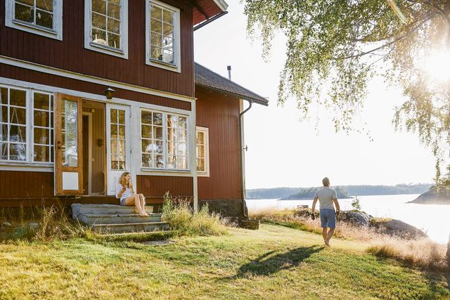 woman sitting on steps at entrance of log cabin while man walking towards lake on sunny day