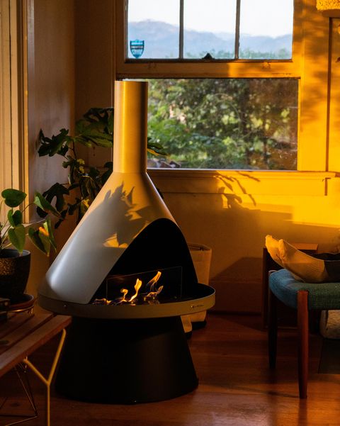 Bitkilerle çevrili bir oturma odasının bir köşesinde Lloyd modern jel yakıtlı şömine ve sandalyede oturan bir kişi