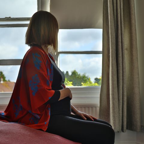 lone pregnant woman sitting on the bed looking out at the window in the morning