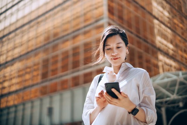 low angle portrait of young asian businesswoman checking emails on smartphone outside office building in financial district with contemporary corporate skyscrapers in background making business connections throughout the city