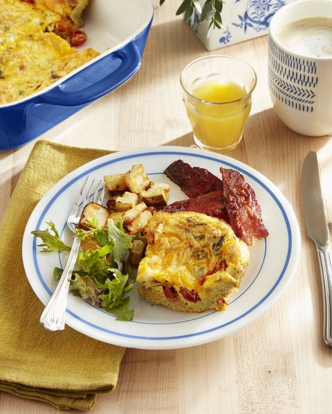 a serving of tomato and fresh basil frittata on a white plate with blue trim alongside a couple pieces of bacon and potatoes and a small salad