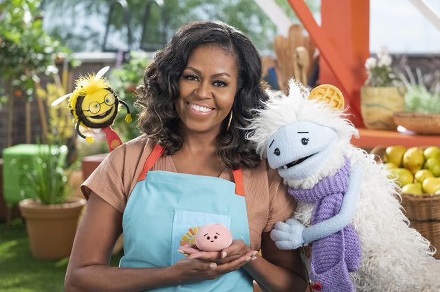 michelle obama, wearing a blue apron, stands in a rooftop garden posing for the camera and holding a pink, round mochi puppet a bee puppet wearing a red tie hovers over her shoulder while a furry white and blue puppet with frozen waffle ears embraces her side