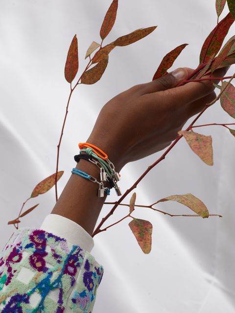 A single hand wearing all four different colors of the Silver Lockit bracelet (orange, green, black, and blue) holds a thin branch with oranging leaves.