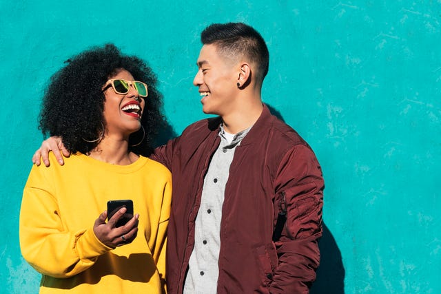 man and woman laughing while using mobile phone while standing against turquoise wall