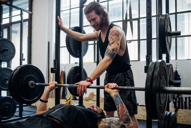 man lying on a weight bench about to bench press with another man standing with his hand on the bar