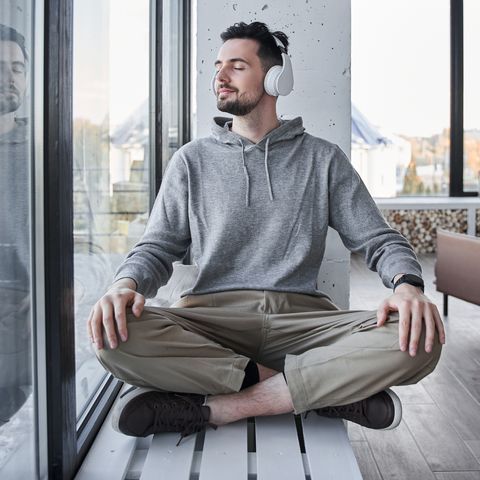 man wearing headphones keeping eyes closed and sitting on windowsill in a lotus position