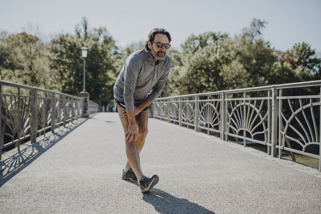 man wearing sunglasses stretching leg while standing on footbridge during sunny day