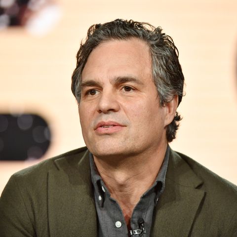 mark ruffalo sits in front of a backdrop featuring a giant hbo logo during a press event