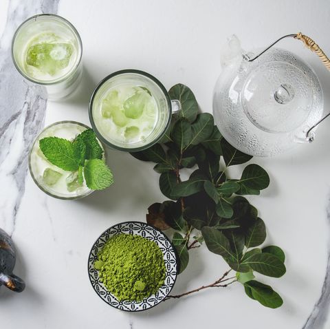 matcha green tea iced latte or cocktail in three different glasses with ice cubes, matcha powder and jug of milk on white marble table, decorated by green branches grey wall at background