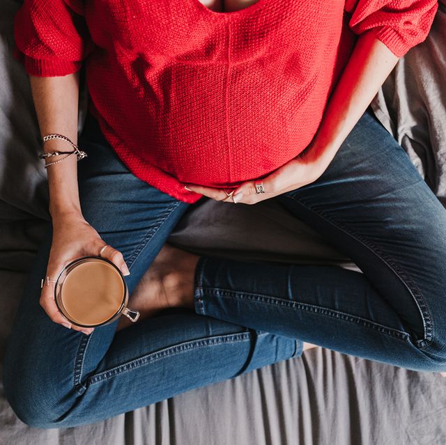 top view of pregnant woman in red sweater holding their baby bump while sitting cross legged in jeans on a bed and holding a mug of coffee
