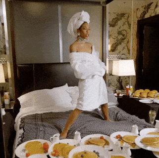 mujer con albornoz baila en la cama en una habitación de hotel de lujo repleta de comida