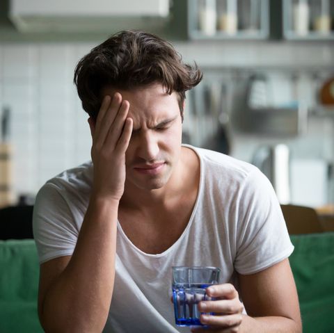 young man suffering from strong headache or migraine sitting with glass of water in the kitchen, millennial guy feeling intoxication and pain touching aching head, morning after hangover concept