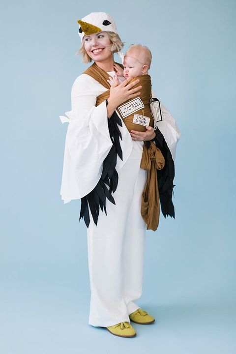 woman in stork costume wearing white with black fringe on the long sleeves, and a stork head with black eyes and the upper half of a bill on her head she is holding a baby in a brown wrap that has signs that read special deliver and handle with care