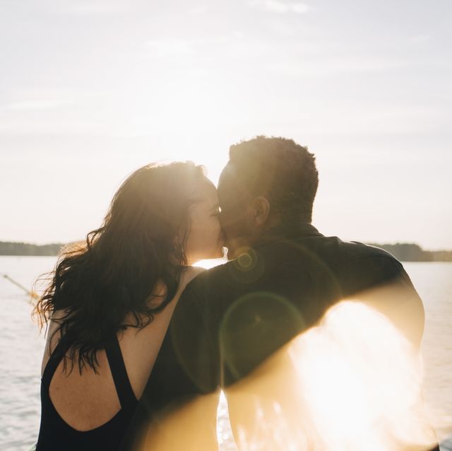 Multi-ethnic couple kissing while sitting against lake during summer